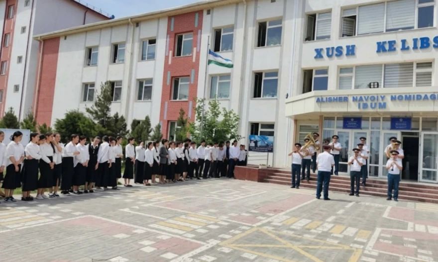 Maktabning bosh binosi: bayroq, orkestr va safga tizilgan oʻquvchilar